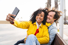 Smiling Woman With Man Taking Selfie Through Mobile Phone Sitting On Wooden Bench