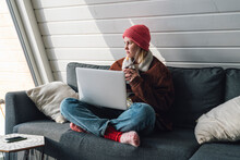 Woman In Warm Clothing Sitting Cross-legged On Sofa With Laptop At Home