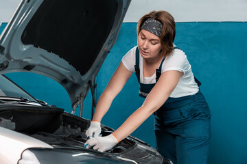 Wall Mural - Young woman mechanic in coveralls and gloves repairs the engine. Mock up with copy space. The concept of women's equality and service in an auto repair shop