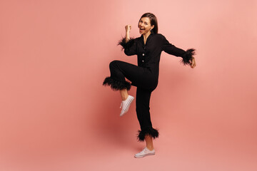 Wall Mural - Full length young caucasian woman clenches fist, bends knee, rejoices in victory on pink background. Brunette wears black silk pajama shirt and pants. Happy morning concept