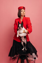 Wall Mural - Confident young caucasian girl with dog hugs beloved pet with love on pink background. Brunette with short haircut wears red jacket and black skirt. Relationship between animals and people, concept
