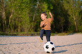 Fototapeta Sport - Kid Kicks Football Ball. Funny Little Boy Playing with Black and White Soccer Ball in Park on Sand