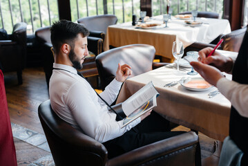 Wall Mural - A young businessman in a fine restaurant examines the menu and makes an order to a young waiter in a stylish apron. Customer service.