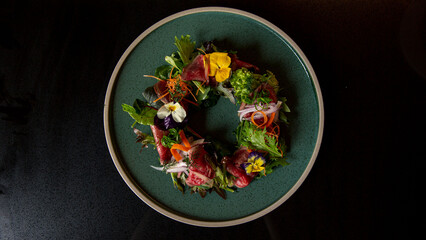 Top view of Tartare beef flower circle salad in the fine dining Japanese restaurant, with a green round plate in black scene reflection table background, raw beef, onion, salad, carrot, flowers