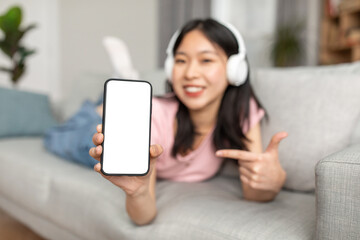 Great musical app. Happy asian lady showing phone empty screen, wearing wireless headphones, lying on sofa at home