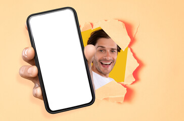 Wall Mural - Cheerful guy showing white empty smartphone screen through torn paper