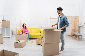 Wall Mural - Cheerful woman holding book near arabian boyfriend and packages at home.