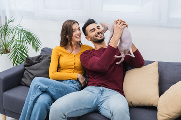 Wall Mural - Positive multiethnic couple looking at sphynx cat on couch.