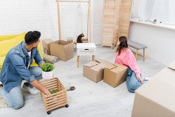 Wall Mural - Side view of multiethnic couple looking at bengal cat near packages in living room.