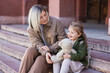 © LIGHTFIELD STUDIOS - blonde woman looking at happy daughter sitting on stairs with teddy bear.