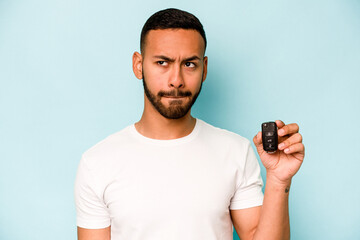 Wall Mural - Young hispanic man holding car keys isolated on blue background confused, feels doubtful and unsure.