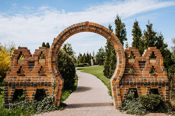 A beautiful round arch made of brown bricks stands on the road at the entrance to the park with plants.