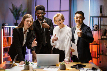 Wall Mural - Portrait of creative multiethnic business team standing together at the table with gadgets and papers and looking at camera. Multiracial business people together at the evening meeting.