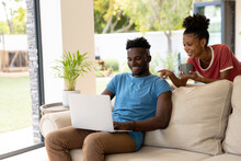 Smiling African American Young Woman Holding Mug Pointing At Laptop Using By Boyfriend On Sofa