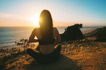 Woman Practicing Yoga And Meditates On The Mountain With Ocean View At Sunset. Young Woman Sitting In Lotus Pose Outdoor In The Nature