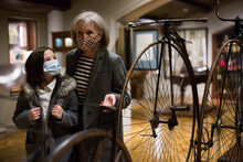Curious Tween Schoolgirl And Friendly Elderly Female Tutor In Face Masks Viewing Vintage Bicycle In Museum Of History Of Technology With Interest. Knowledge Concept. New Normal In Coronavirus Pandemic