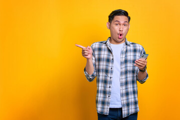 Shocked young Asian man in plaid shirt holding mobile phone and pointing fingers aside at copy space isolated on yellow background