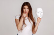 © khosrork - Portrait of dark haired woman pointing finger on you, holding white mask with unknown face, blaming you in duplicity, wearing white T-shirt. Indoor studio shot isolated on gray background.