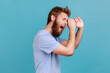 © khosrork - Side view of wondered shocked bearded man making monocular gesture with hands and looking amazed at side space, keeps mouth opened. Indoor studio shot isolated on blue background.