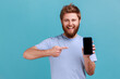 © khosrork - Portrait of smiling happy bearded man pointing finger at smartphone with black blank screen, copy space for advertisement or promotion. Indoor studio shot isolated on blue background.