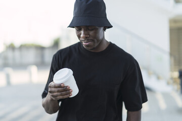 Wall Mural - Young cool black male in a street style outfit walking with a paper white coffee cup