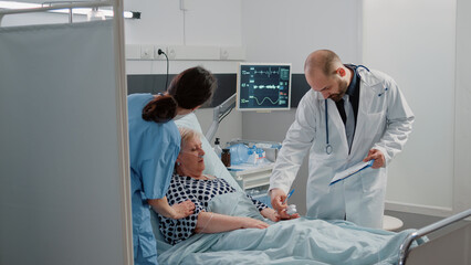 Wall Mural - Ill patient discussing disease and treatment with doctor in hospital ward. Retired woman with nasal oxygen tube and IV drip bag talking to medic at checkup visit while nurse giving support