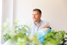 Business Portrait Of Environmental Engineer, Ecologist, Garden Architect Sitting At His Work Table Holding A Globe In His Hands And Surrounded By Plants