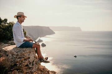 Digital nomad, man in the hat, a businessman with a laptop sits on the rocks by the sea during sunset, makes a business transaction online from a distance. Remote work on vacation.