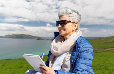 Sticker - travel, tourism and vacation concept - happy senior woman writing to notebook over atlantic ocean coast in ireland background