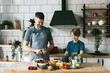 © klavdiyav - family dad young man and son teenage boy cook vegetable salad in kitchen and spend quality time together, father and son talking and cooking vegetarian food and doing chores, 8 march and mothers day
