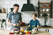 © klavdiyav - family dad young man and son teenage boy cook vegetable salad in kitchen and spend quality time together, father and son talking and cooking vegetarian food and doing chores, 8 march and mothers day