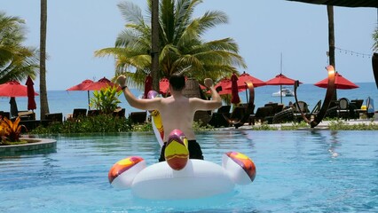 Wall Mural - Happy man have fun ride dance on giant inflatable unicorn in swimming pool in luxury hotel, enjoy holidays or vacations on sunny summer day. Funny unknown adult male tourist in resort. Back view.