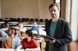 © Seventyfour - Waist up portrait of male college professor looking at camera in classroom hall and holding book, copy space
