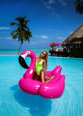 Beautiful sexy tanned woman on pink flamingo in pool. Young glamour girl in swimsuit on Maldives island. Perfect body bikini model in luxury resort on Maldives. Luxury travel. Summertime.