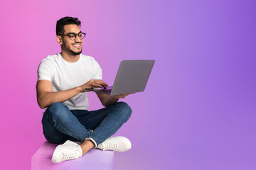 Aufkleber - Full length of millennial Arab man in glasses using laptop for work or studies in neon light, copy space