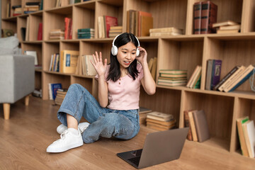 Poster - Young korean lady using laptop for online communication, wearing headphones and waving at webcam