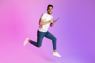 Poster - Happy young Arab man with smartphone jumping up in neon light, copy space. Cool new app, website or advertisement