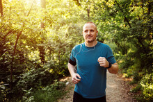 Smiling Male With In-ear Headphones Jogging In Park. Mid Adult Man Is Listening To Music During Summer. He Is Wearing Sports Clothing. Shot Of A Young Man Going For A Morning Run.