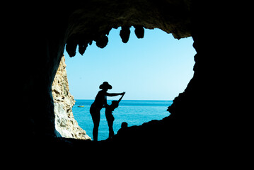 Poster - Silhouette of a female with a kid standing at the entrance of a rocky cave over a sunny blue water