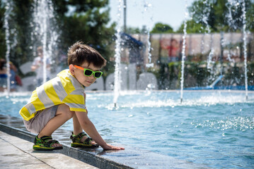 Wall Mural - Little boy plays in the square near pool with water jets in the fountain at sunny summer day. Active summer leisure for kids in the city