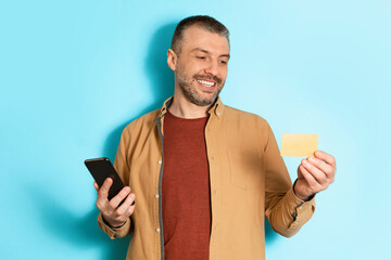 Poster - Cheerful Man Holding Credit Card And Phone Over Blue Background