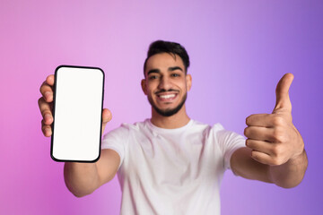 Poster - Happy yougn Arab guy holding cellphone with empty white screen and showing thumb up gesture in neon light, mockup