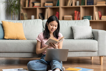Poster - Happy asian lady studying online from home, reading book and using laptop at home, sitting on floor near sofa