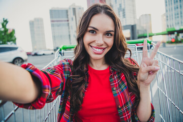 Wall Mural - Self-portrait of beautiful trendy cheerful carefree girl sitting in cart having fun on parking showing v-sign outdoors