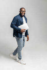 Cheerful African-American man carrying laptop computer isolated on grey. Happy black male entrepreneur with laptop, male student using portative gadget, computer. Vertical photo