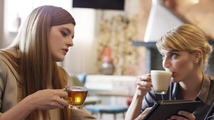 Aufkleber - Two businesswomen talking, drinking herbal tea and coffee while brainstorming in a meeting using a tablet in a cafe. Happy businesswomen collaborating on a marketing project using a tablet.
