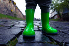 A Small Child In Green Rubber Boots Stands On A Paved Road.