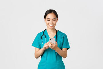 Wall Mural - Covid-19, healthcare workers and preventing virus concept. Smiling happy beautiful asian female intern, doctor making phone call, looking at mobile phone screen pleased, messaging or using app