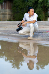 Poster - Asian man holds a big black mirrorless camera medium format type on cement asphalt ground in front of water reflex.