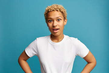 Wall Mural - Funny surprised woman of African ethnicity wearing white t-shirt and nose ring, standing with mouth opened with astonishment against blue background after learning unbelievable, shocking news